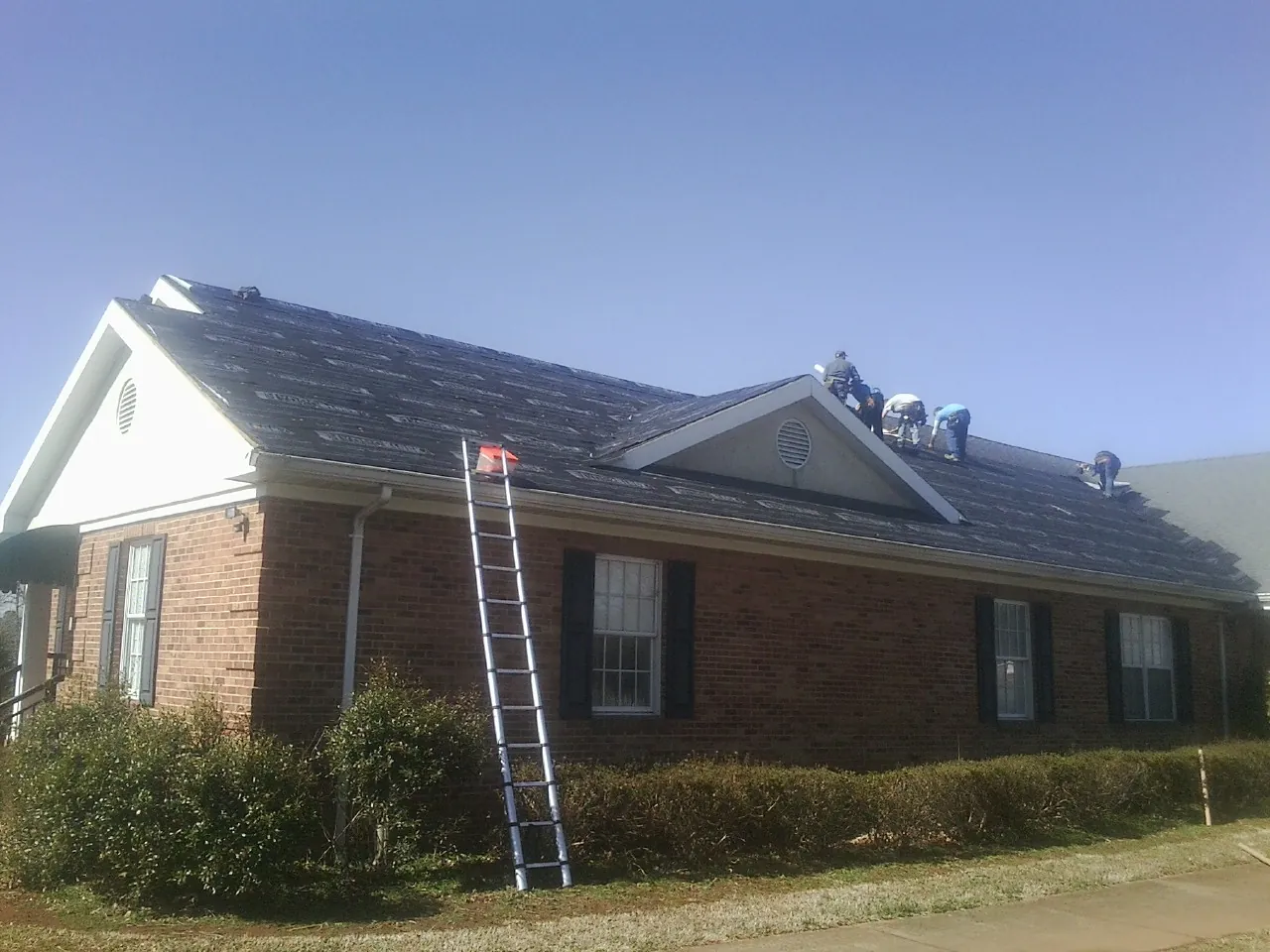 Crew tearing shingles off a brick ranch roof with a ladder set — Metal Roof Repair work by our crew in Hadley